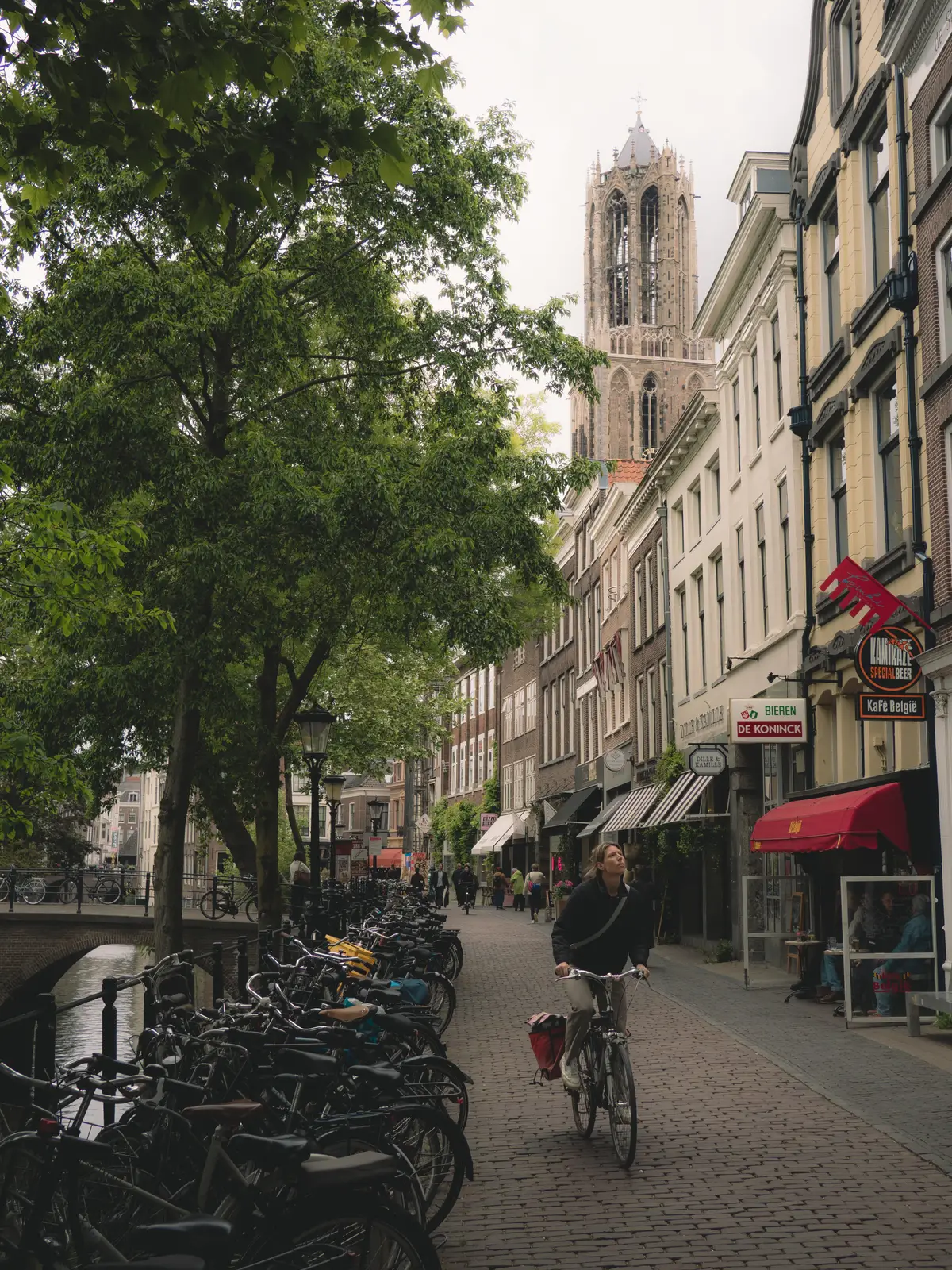 A woman cycling down a cobblestone canal-side street lined with parked bikes, the Dom Tower rising in the background